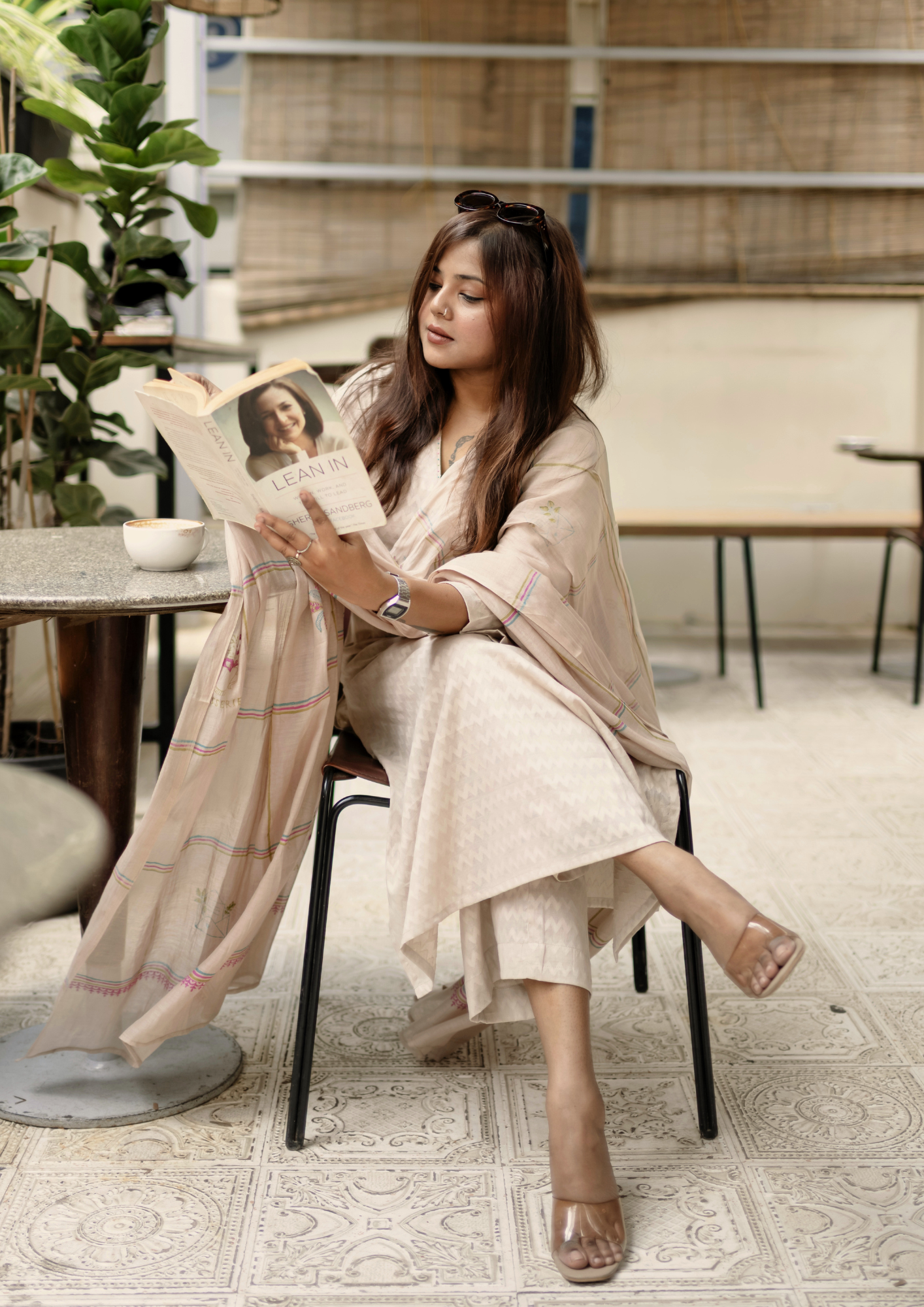An Indian Woman wearing a light beige colour pure cotton Indian suit set, she is sitting in a cafe reading a book and sipping on coffee